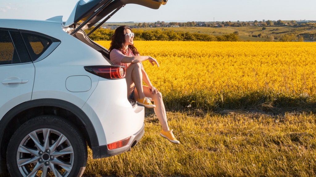 Eine lächelnde, hübsche Frau sitzt bei Sonnenuntergang im Kofferraum eines SUVs und blickt auf ein Ackerfeld (Roadtrip-Konzept).