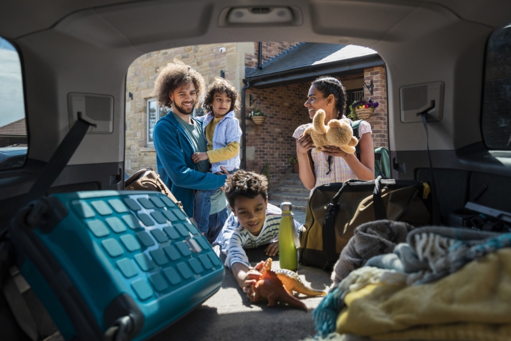 Eine Halbtotale aus dem Inneren eines Autos zeigt eine Familie, die den Kofferraum auspackt, ihre Koffer und das Spielzeug der Kinder holt, als sie in der Lodge ankommen, in der sie ihren Urlaub im Nordosten Englands verbringen. Der Vater trägt einen seiner Söhne.